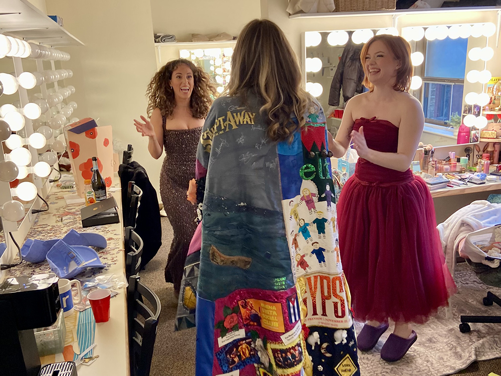 In a dressing room, Mia Geriachis and Taylor Sage Evans jump for joy. Between them, Martignetti's back is visible in the Robe.