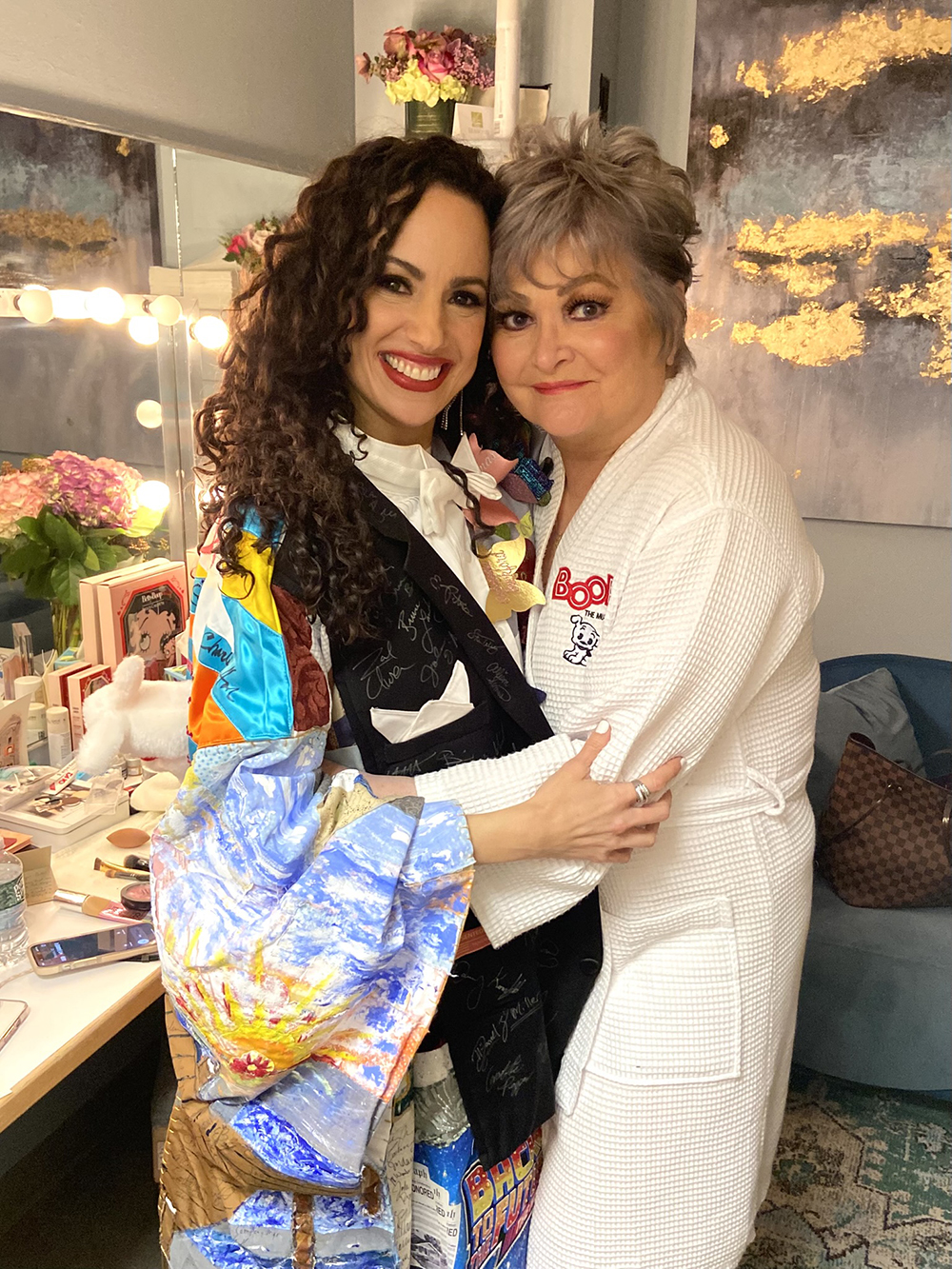 Lafarga, smiling and wearing the Robe, hugs Faith Prince in a dressing room. Prince is wearing a 'Boop! The Musical' branded bathrobe. 
