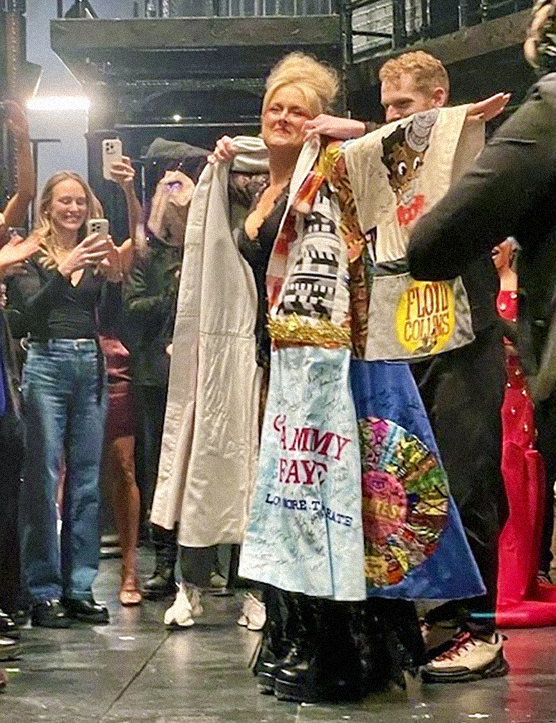 DeLaney Westfall smiles onstage as Max Chernin puts the Robe onto her, as excited onlookers applaud and take pictures.