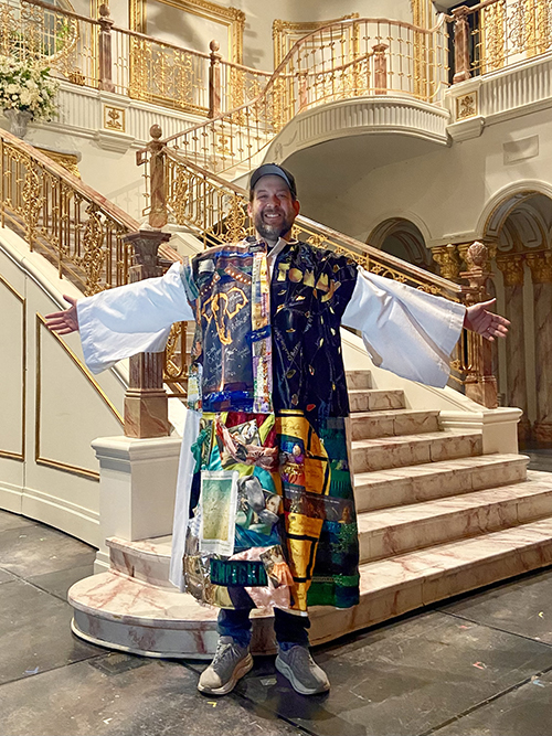 Andrew Kober stands with his arms outstretched wearing the Robe, at the foot of the marble staircase on the set of the stage.