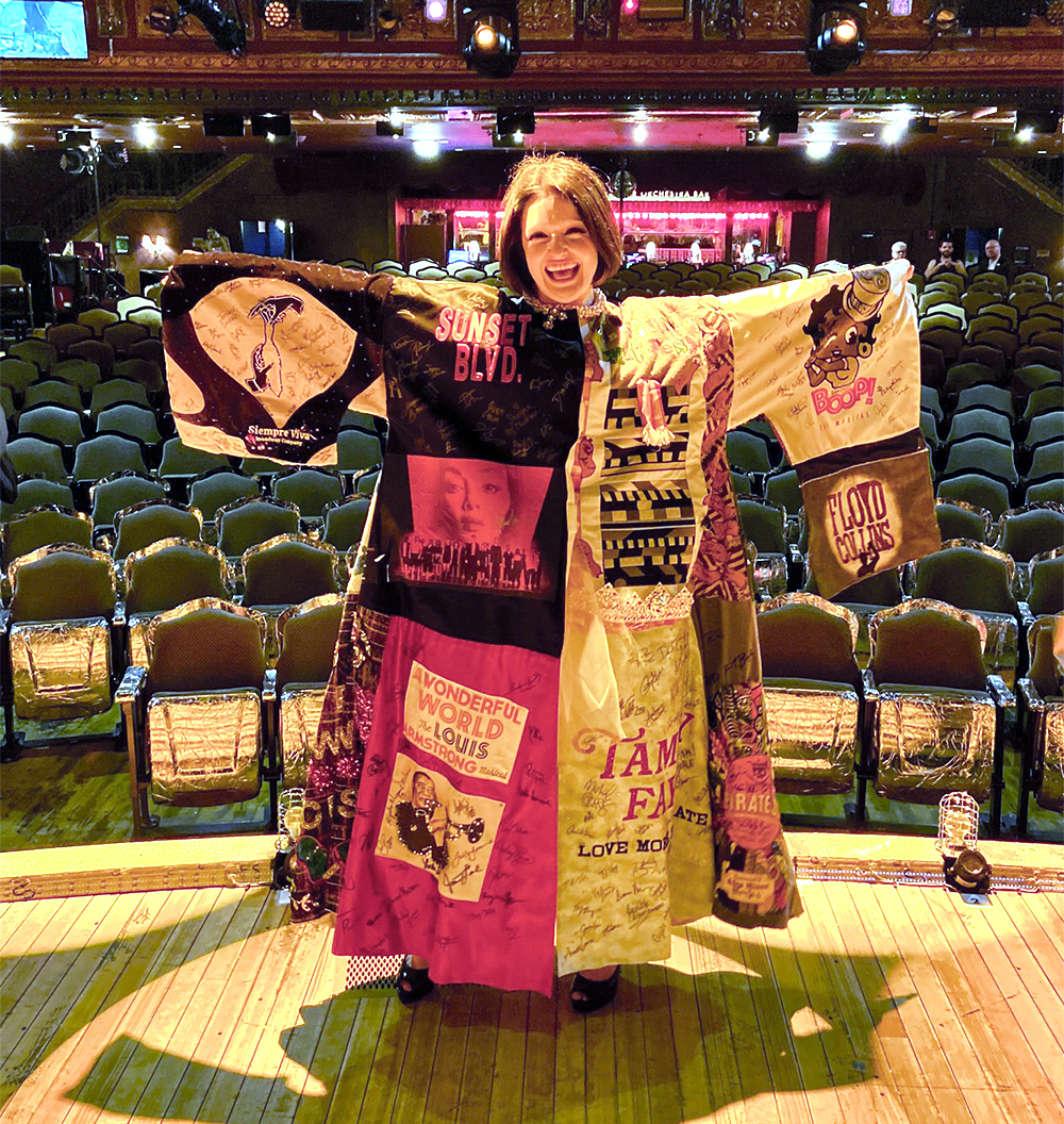 Larkin Reilly smiles onstage in the Robe, arms outstretched. There are house seats visible behind her. The lighting is yellow and somewhat eerie.