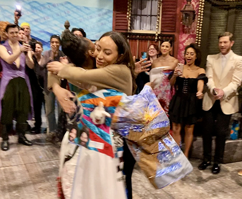 Hines embraces Khori Michelle Petinaud, who is wearing the Robe. Several people stand behind them looking excited, the set of the musical partially visible..