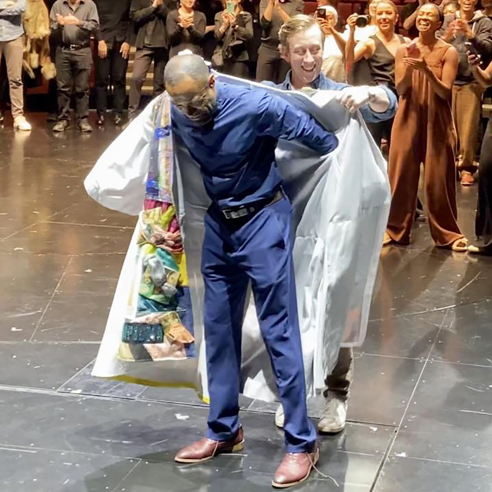 Onstage surrounded by excited people taking photos and video, Ryan Breslin holds the Robe for Alan Wiggins to don.